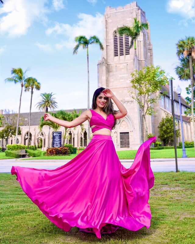 Roopsi Bansal twirling in a magenta satin crop top and full skirt, in front of the Gothic bell tower of the Church of Bethesda-by-the-Sea, Florida