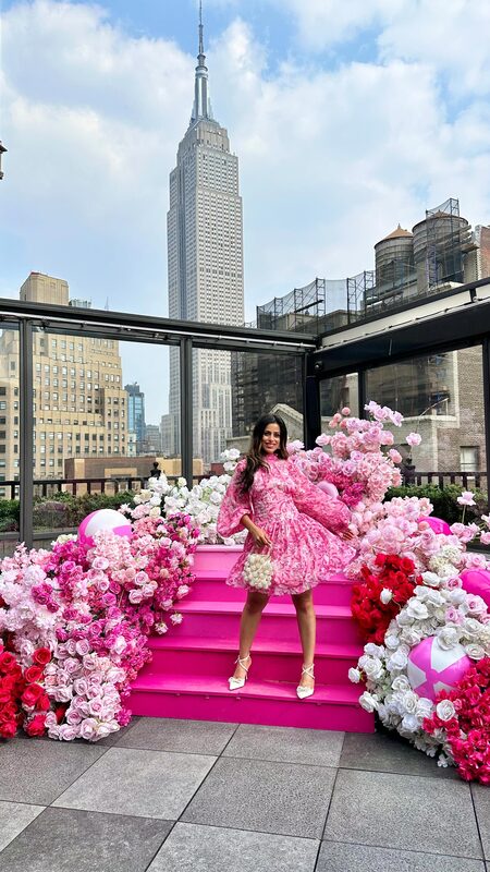 Roopsi Bansal in a pink floral dress standing on a hot pink flower-covered staircase at Magic Hour rooftop, with the Empire State Building behind her