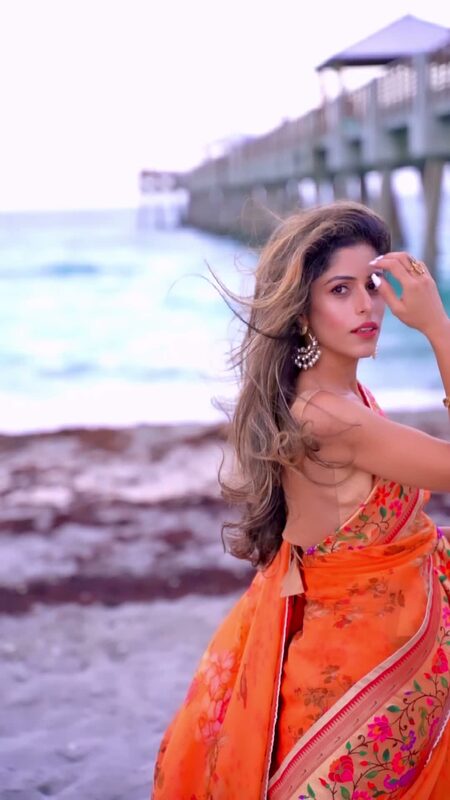 Roopsi Bansal in a flowing fire-orange organza saree with a floral border, hair blowing in the wind, standing beneath Juno Beach Pier with waves behind her