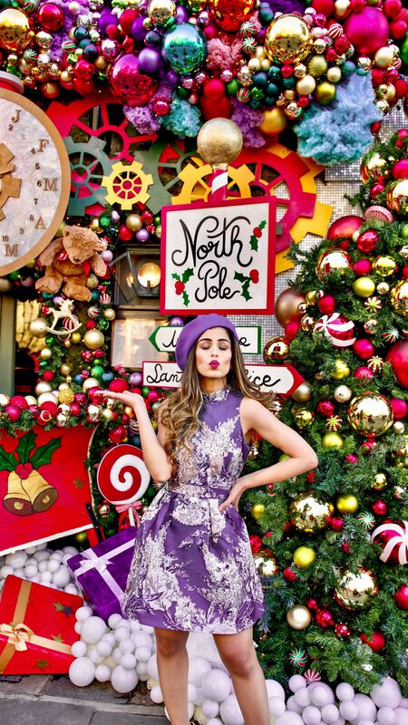 Roopsi Bansal in a lavender floral dress and purple beret, blowing a kiss beneath a 'North Pole' signpost surrounded by ornaments and a decorated Christmas tree