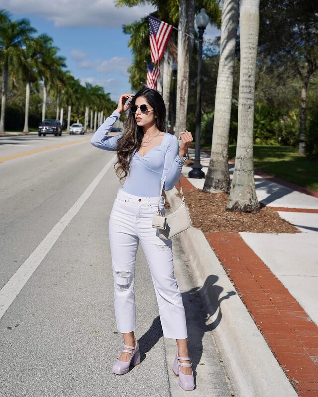 Roopsi Bansal in a light blue silky thong bodysuit and high-waisted white ripped jeans, standing on a palm-lined street with American flags overhead