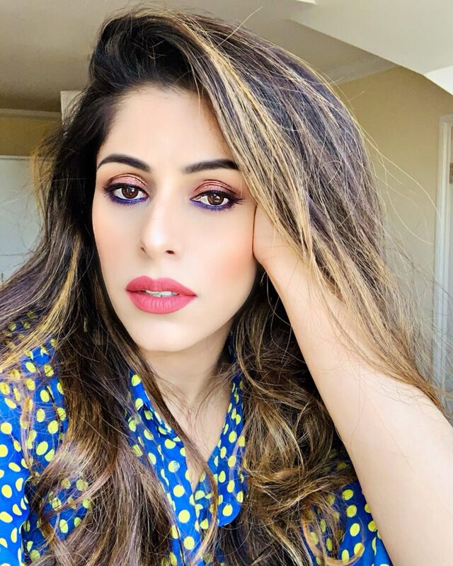 A close-up selfie with rose-gold and purple eyeshadow and a coral matte lipstick, wearing a blue and yellow polka-dot top