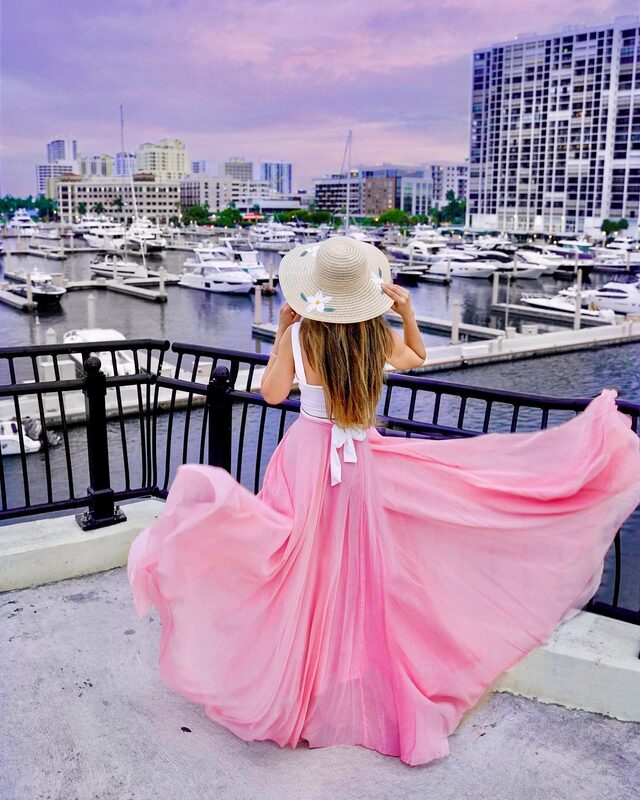 Roopsi Bansal from behind in a flowing pink chiffon maxi skirt and straw hat, leaning on a railing overlooking a yacht marina at dusk