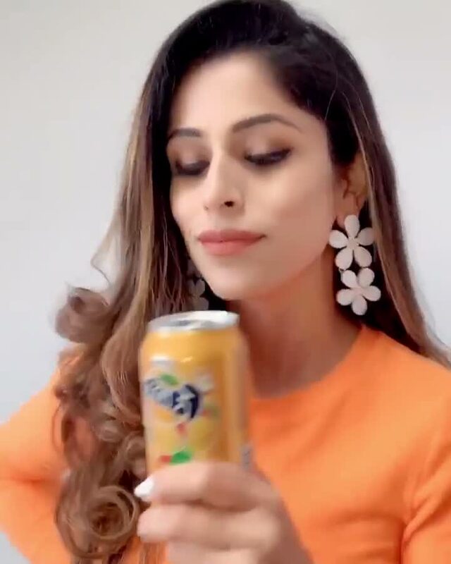 Roopsi Bansal in a bright orange top holding a can of orange soda, matching her outfit to her drink for an outfit-change challenge video