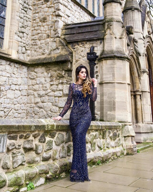 Roopsi Bansal in a navy sequin mermaid gown with long lace sleeves, standing in front of a Gothic stone church facade