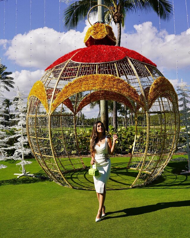 Roopsi Bansal in a white sleeveless dress with a floral applique, holding a neon green handbag, standing inside a giant red and gold ornament sculpture with palm trees behind her
