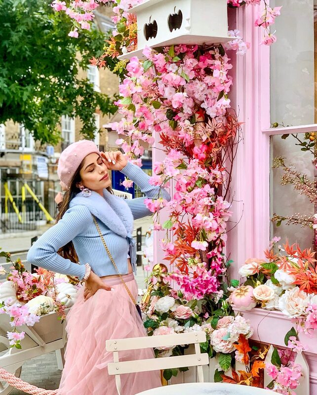 Roopsi Bansal in a pink beret, blue faux-fur trimmed sweater, and pink tulle skirt, reaching up to a wall covered in pink cherry-blossom style flowers