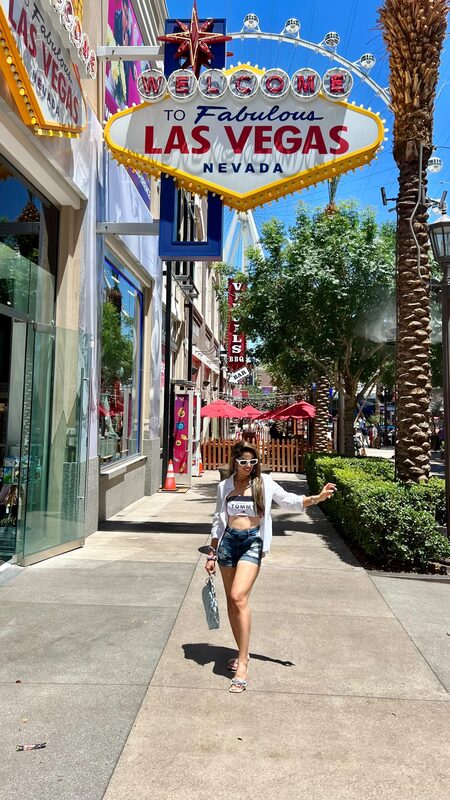 Roopsi Bansal in denim shorts and a white shirt, walking beneath the Welcome to Fabulous Las Vegas Fremont Street sign
