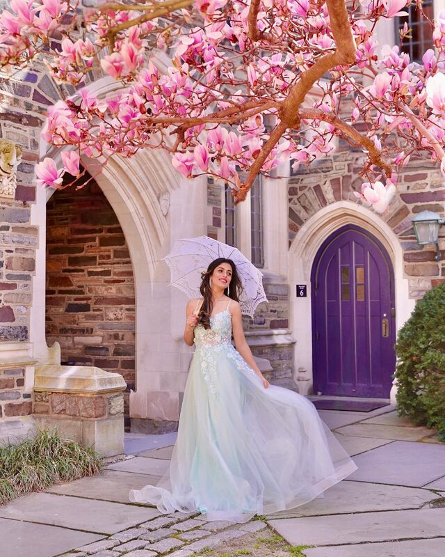 Roopsi Bansal in a mint lace ballgown holding a white lace parasol beneath a blooming magnolia tree at a purple Gothic door