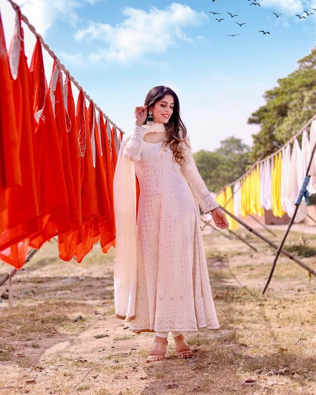 Roopsi Bansal in a white chikankari embroidered anarkali suit, standing between rows of orange, yellow, and white dupattas hung out to dry