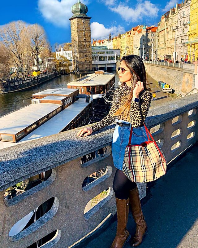 Leaning on a stone bridge railing over the river in Prague, wearing a leopard-print turtleneck, denim skirt with a Gucci belt, tall brown boots, and carrying a large Burberry check tote