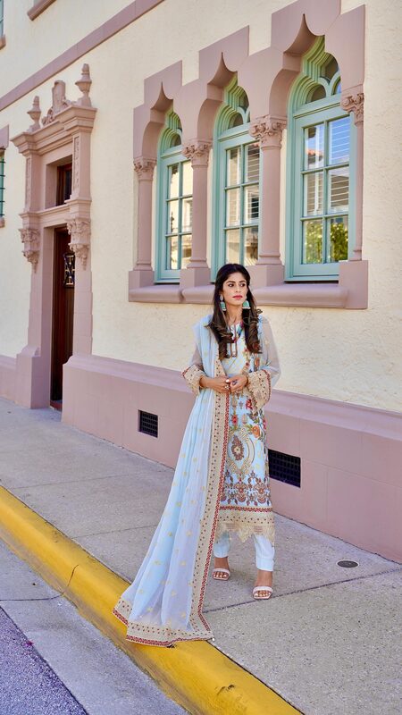 Roopsi Bansal in a pale blue embroidered salwar suit from Khas and green tasseled earrings from Jaipur Rose, standing in front of a Mediterranean Revival building with arched green-shuttered windows