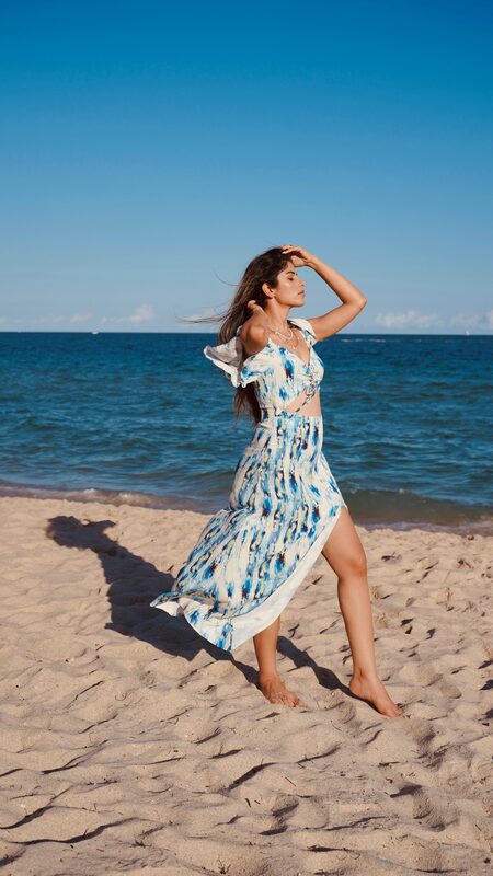 Roopsi Bansal in a blue and white tie-dye cutout maxi dress, hair blowing in the wind, standing barefoot on the sand at the water's edge