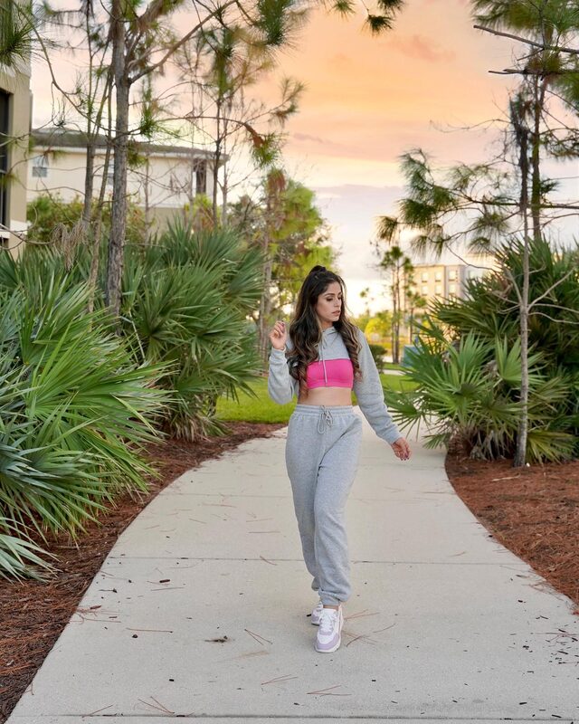 Roopsi Bansal in a gray cropped hoodie over a pink sports bra and matching gray sweatpants, walking down a pine-lined path at golden hour