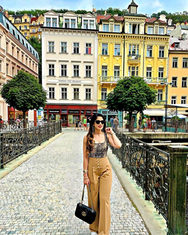 Roopsi Bansal in a camel wide-leg trouser set standing on a bridge in front of the colorful buildings of Karlovy Vary, Czech Republic