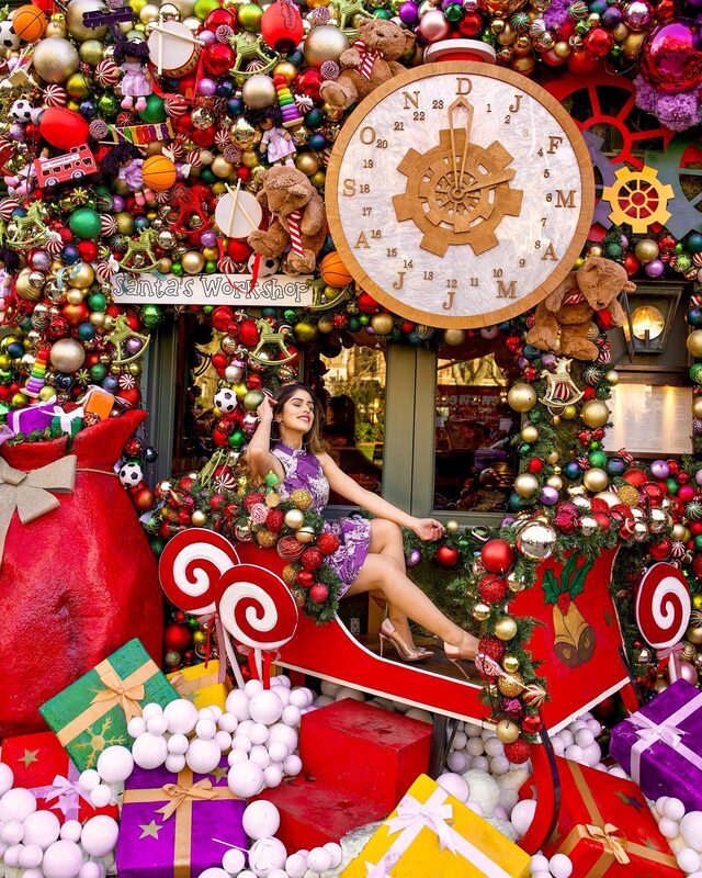 Roopsi Bansal in a purple floral dress, sitting in a red sleigh display under a 'Santa's Workshop' sign surrounded by giant ornaments, teddy bears and a wooden toy clock