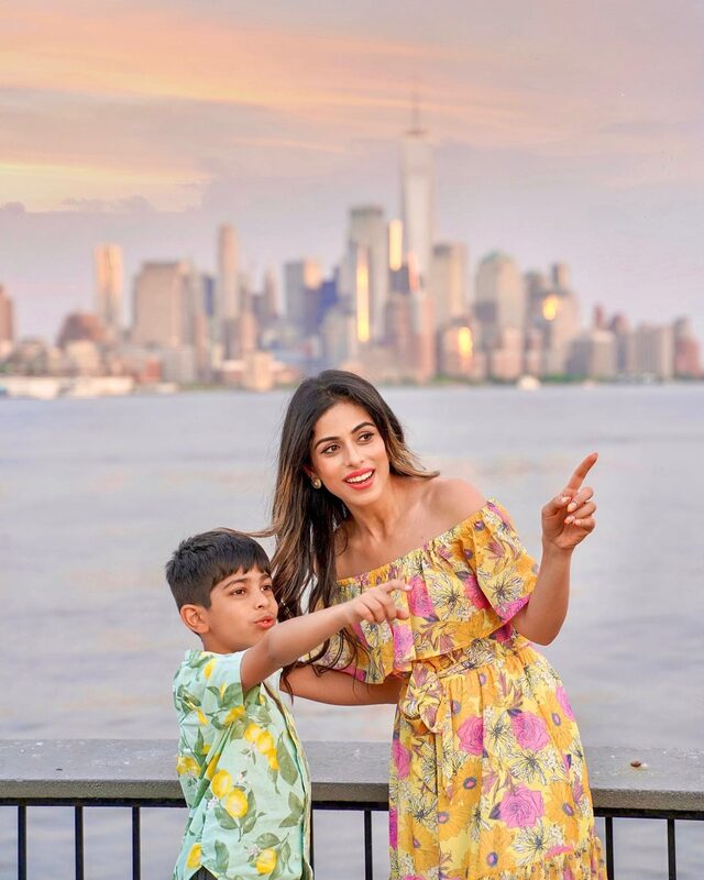 Roopsi Bansal in a yellow floral dress pointing at the sky with her young son, the lower Manhattan skyline glowing at sunset behind them