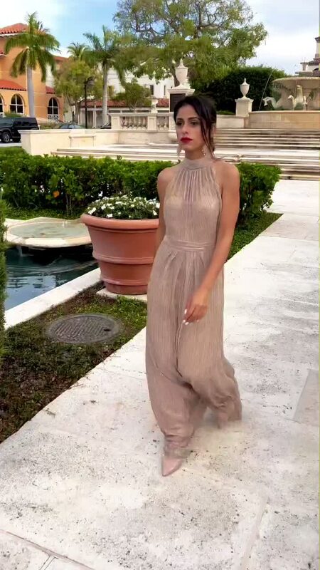 Roopsi Bansal in a metallic champagne gold pleated halter gown from Adrianna Papell, standing on a terrace beside a stone fountain courtyard