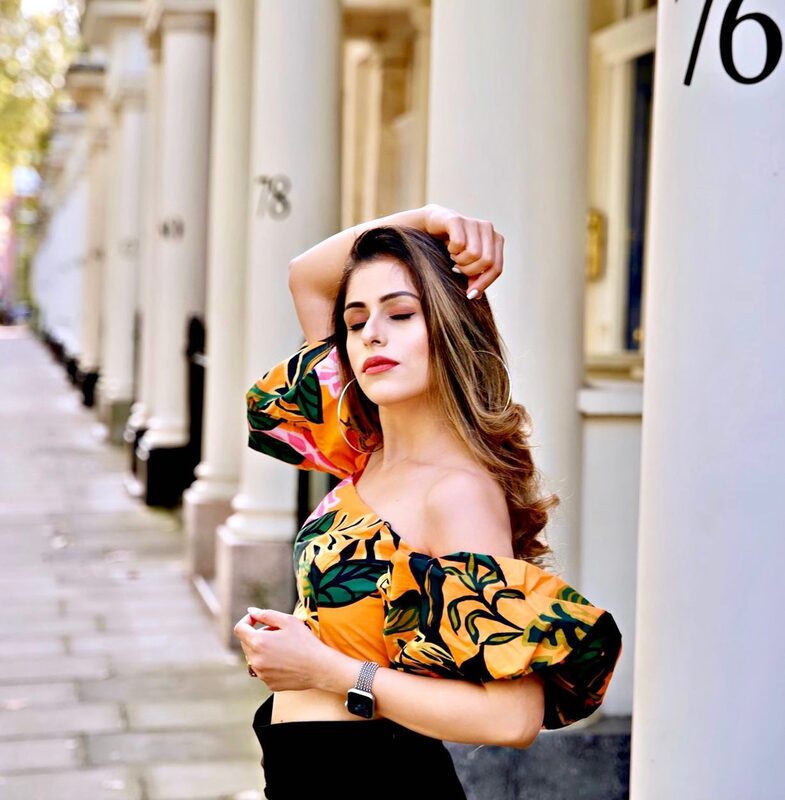 Roopsi Bansal in an orange and green tropical print off-shoulder top, posing against a row of white columns on a London street