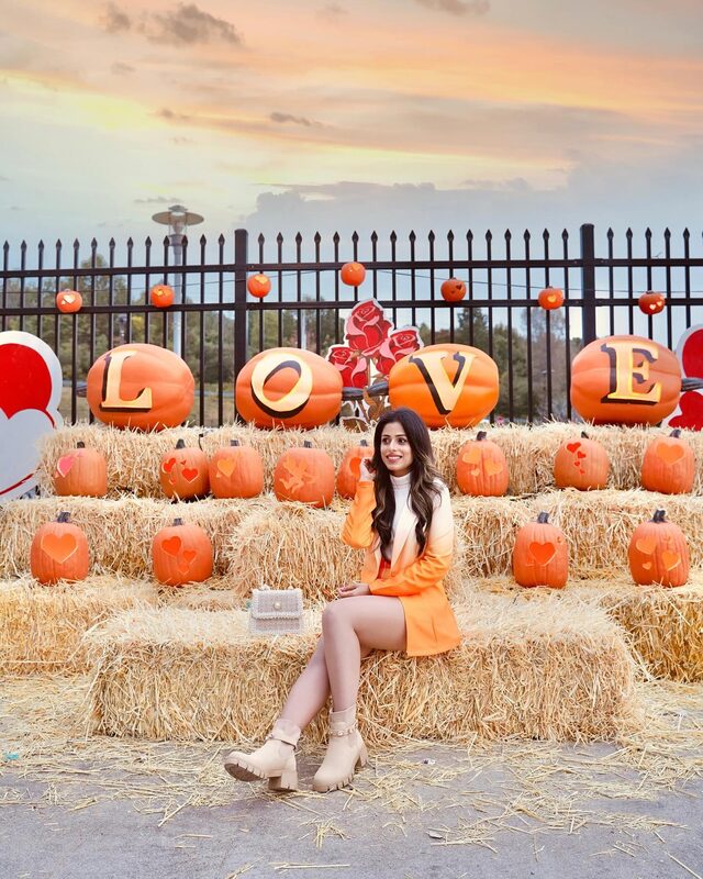Roopsi Bansal in an orange blazer and shorts set from Shein sitting on hay bales in front of a giant pumpkin 'LOVE' display at Pumpkin World