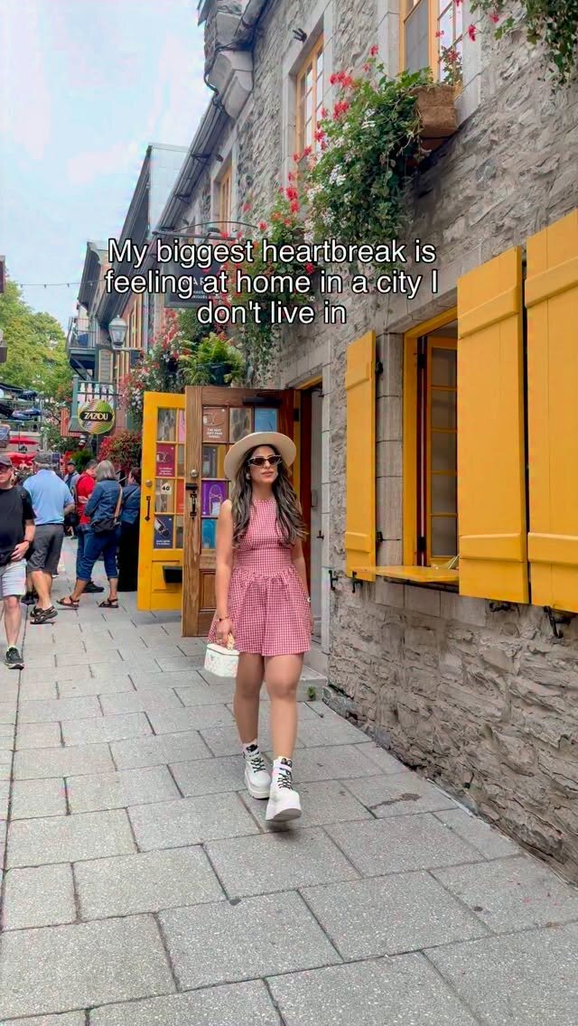 Roopsi Bansal walking down a cobblestone street in Québec City in a pink gingham dress and straw hat, past stone buildings with yellow shutters and flower boxes