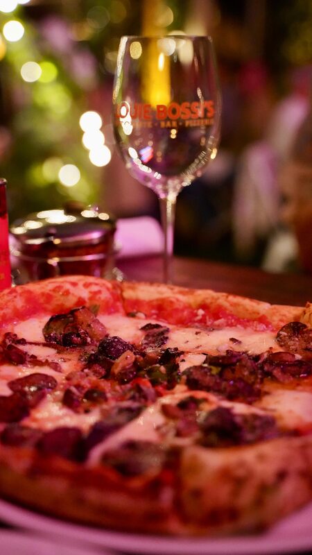 A close-up of a topped pizza with a wine glass branded Louie Bossi's in the background, string lights glowing behind