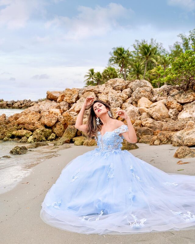 Roopsi Bansal in a sky-blue off-shoulder floral ball gown, seated among rocks on the beach at Dubois Park, Florida