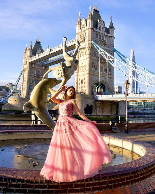 Roopsi Bansal in a pink tulle princess gown, standing in front of the Girl with a Dolphin sculpture with Tower Bridge behind her