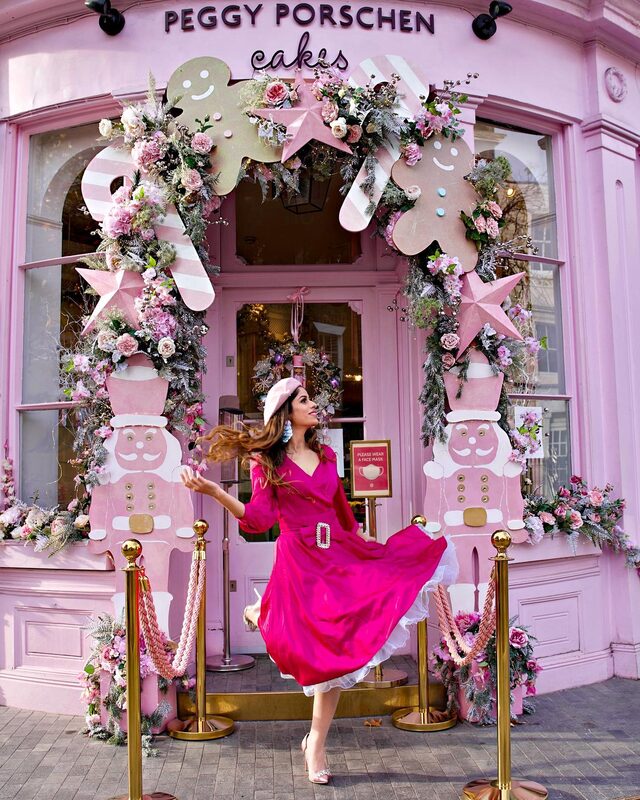 Roopsi Bansal in a hot-pink wrap dress and matching beret, twirling in front of the Peggy Porschen Cakes shopfront decorated for Christmas