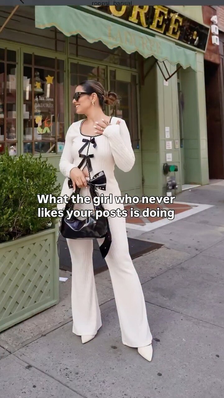 Roopsi Bansal in a cream ribbed knit set with black bow trim, black sunglasses, her hair in a bun, and a black bag, smiling on a New York sidewalk outside Ladurée