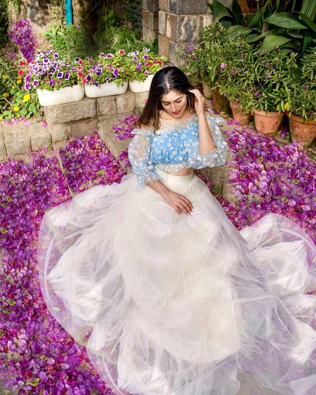 Roopsi Bansal in a pale blue daisy-print off-shoulder top and cream tulle skirt, sitting on stone steps scattered with fallen purple bougainvillea petals