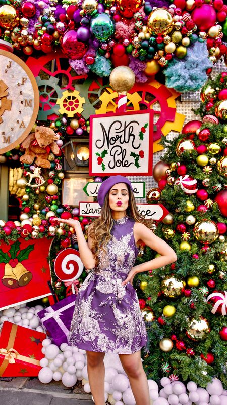Roopsi Bansal in a metallic purple dress and beret in front of a North Pole themed Christmas display with ornaments, a giant clock, and candy decorations