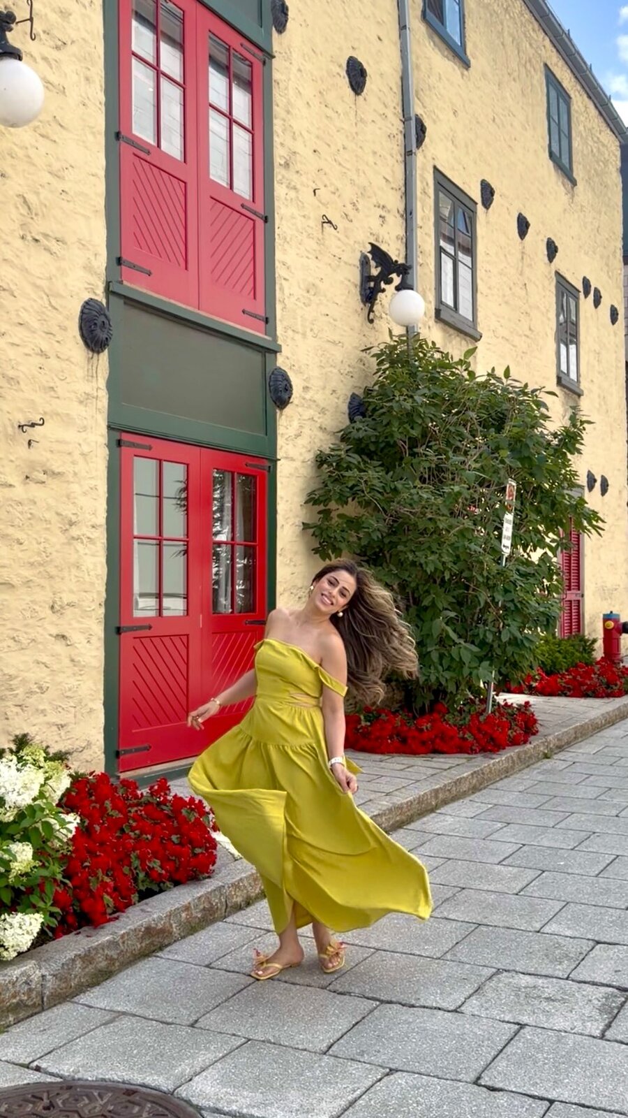 Roopsi Bansal twirling in a yellow off-shoulder dress on a cobblestone street in front of a yellow building with red doors and red flowers in Old Quebec City