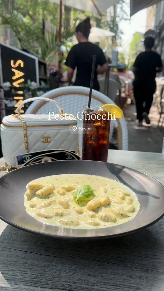 A plate of pesto gnocchi garnished with basil on a restaurant patio table at Savini, with a Chanel bag and sunglasses in the background