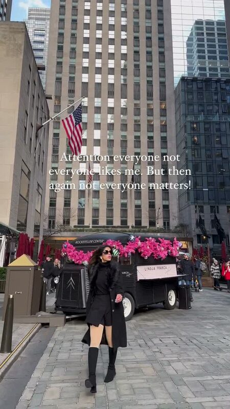 Roopsi Bansal in an all-black outfit walking past a vintage café truck decorated with pink garland at Rockefeller Center