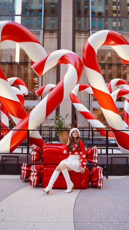 Roopsi Bansal in a Santa sweater at the corner of 6th Avenue and 50th Street in New York beside a Christmas candy decoration