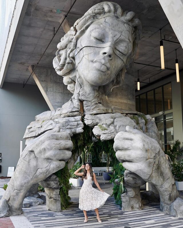 Roopsi Bansal in a white lace midi dress from Dress the Population, twirling beneath the towering stone Thrive sculpture in Fort Lauderdale