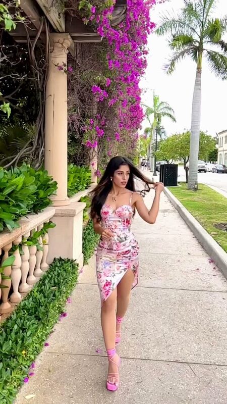 Roopsi Bansal in a pink floral print split-thigh ruched cami dress from SHEIN and pink strappy heels, walking past a colonnade draped in magenta bougainvillea in West Palm Beach