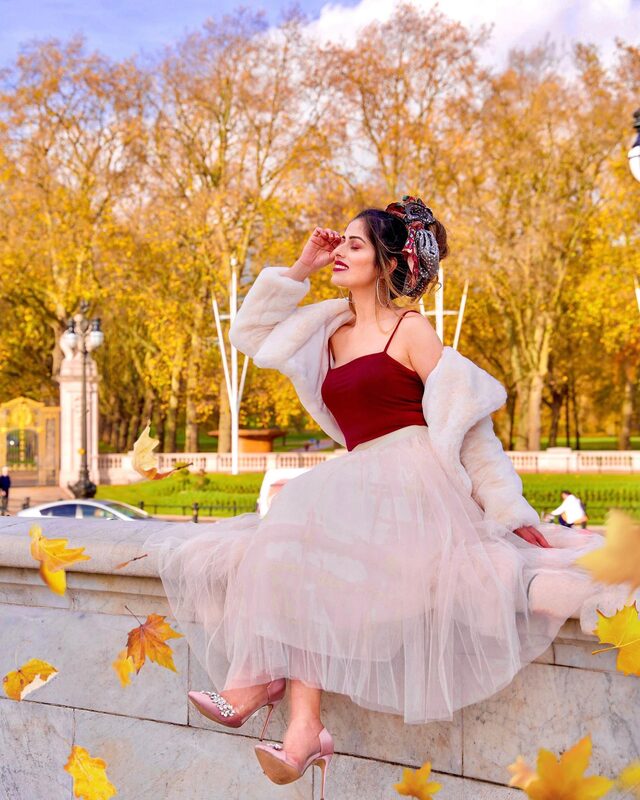 Roopsi Bansal in a cream faux-fur jacket, burgundy cami and blush tulle skirt, sitting on a wall in front of Buckingham Palace's autumn gardens