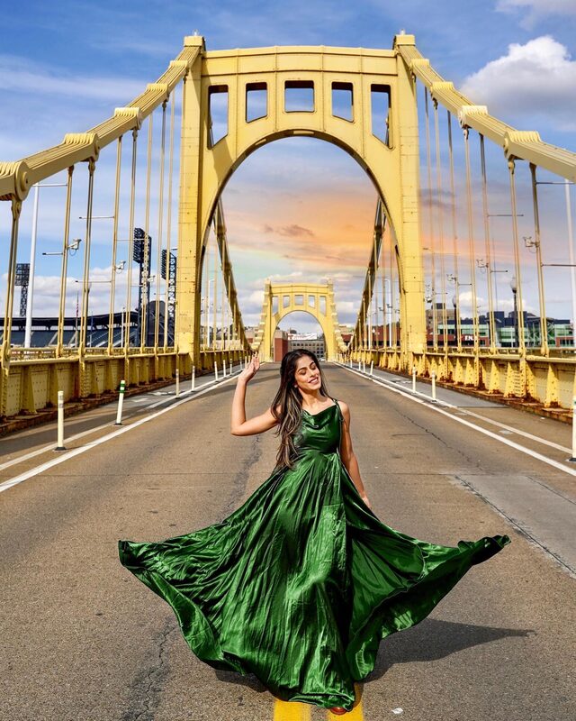 Roopsi Bansal in a flowing emerald green satin gown, twirling on the yellow Roberto Clemente Bridge in Pittsburgh, Pennsylvania, with the city skyline behind her