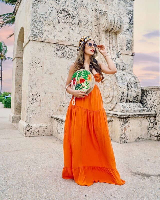 Roopsi Bansal in a tiered orange maxi dress, leopard-print bucket hat, and round sunglasses, holding an embroidered straw bag in front of a carved coral-stone fountain