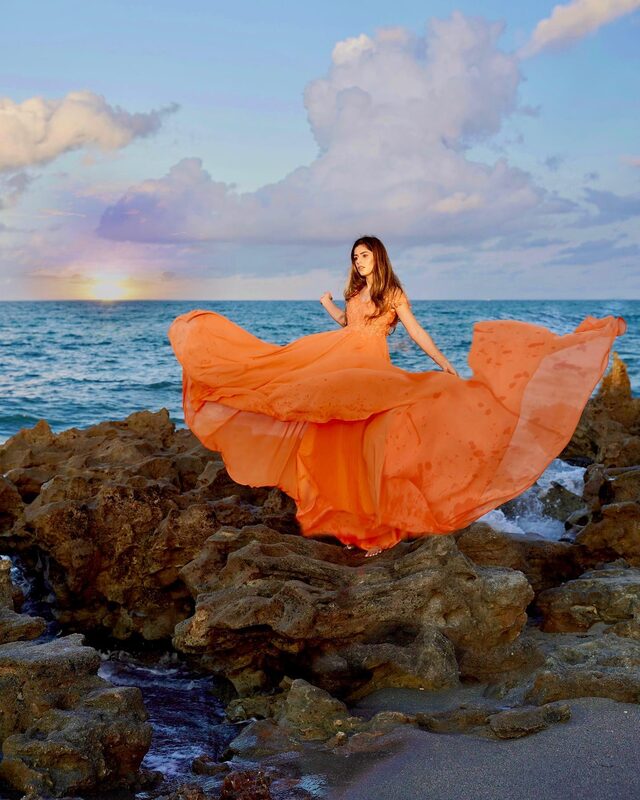 Roopsi Bansal in a flowing orange chiffon gown, standing barefoot on the rocky shore of Coral Cove Park at sunset with the ocean and sun behind her