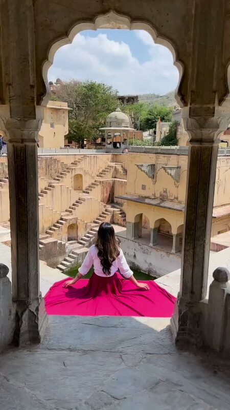 Roopsi Bansal in a pink Chicwish skirt framed by a scalloped stone archway overlooking Panna Meena ka Kund stepwell in Jaipur, India