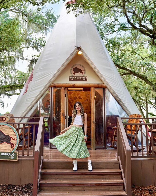Roopsi Bansal in a white crop top and green windowpane check midi skirt, twirling on the porch steps of a canvas glamping tent called Roaming Buffalo, oak trees overhead