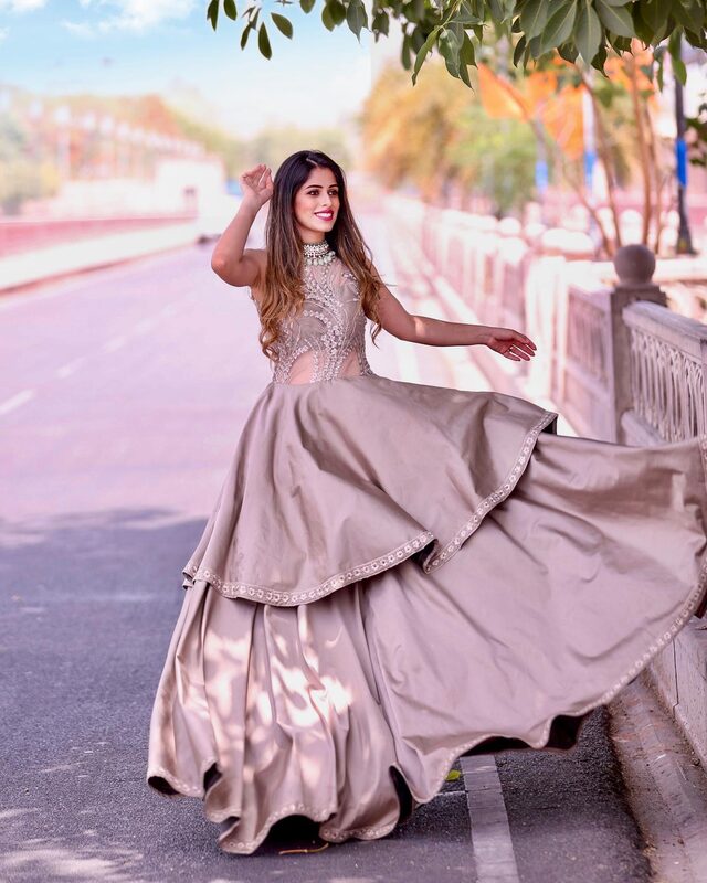 Roopsi Bansal twirling in a pastel taupe embellished ballgown with a beaded statement necklace, on a wide tree-lined city boulevard
