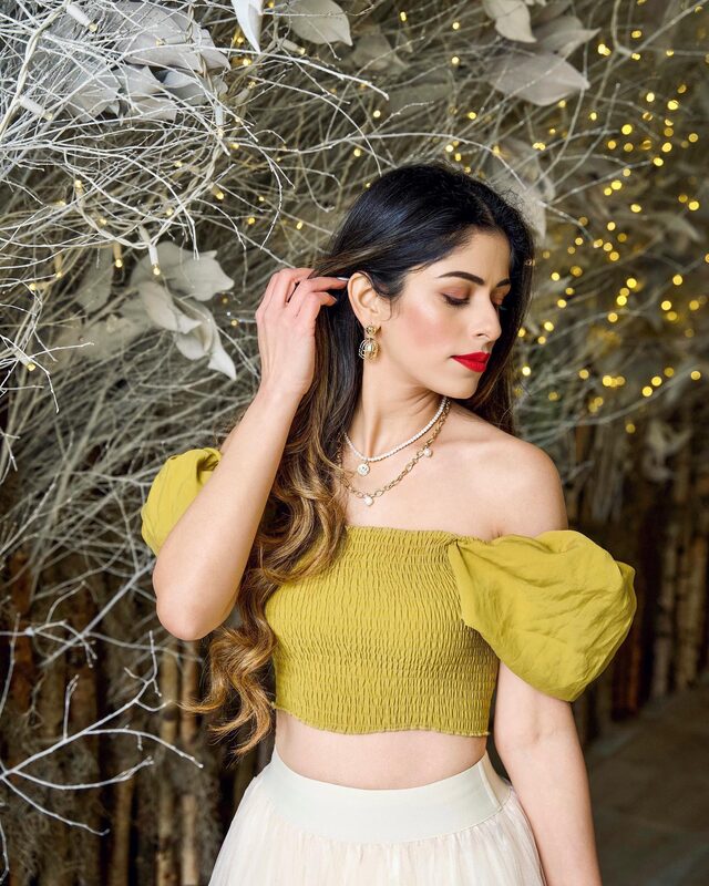 Roopsi Bansal in a mustard off-shoulder top with layered pearl necklaces and gold drop earrings, standing under frosted branches strung with fairy lights