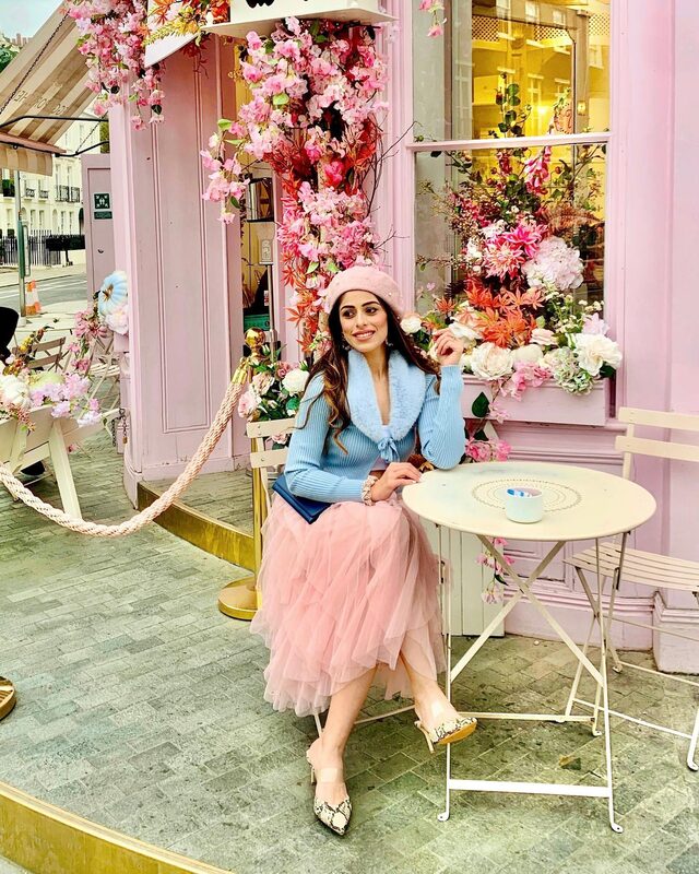 Roopsi Bansal in a pink tulle skirt and pearl beret seated at an outdoor café table at the flower-covered Peggy Porschen café in London