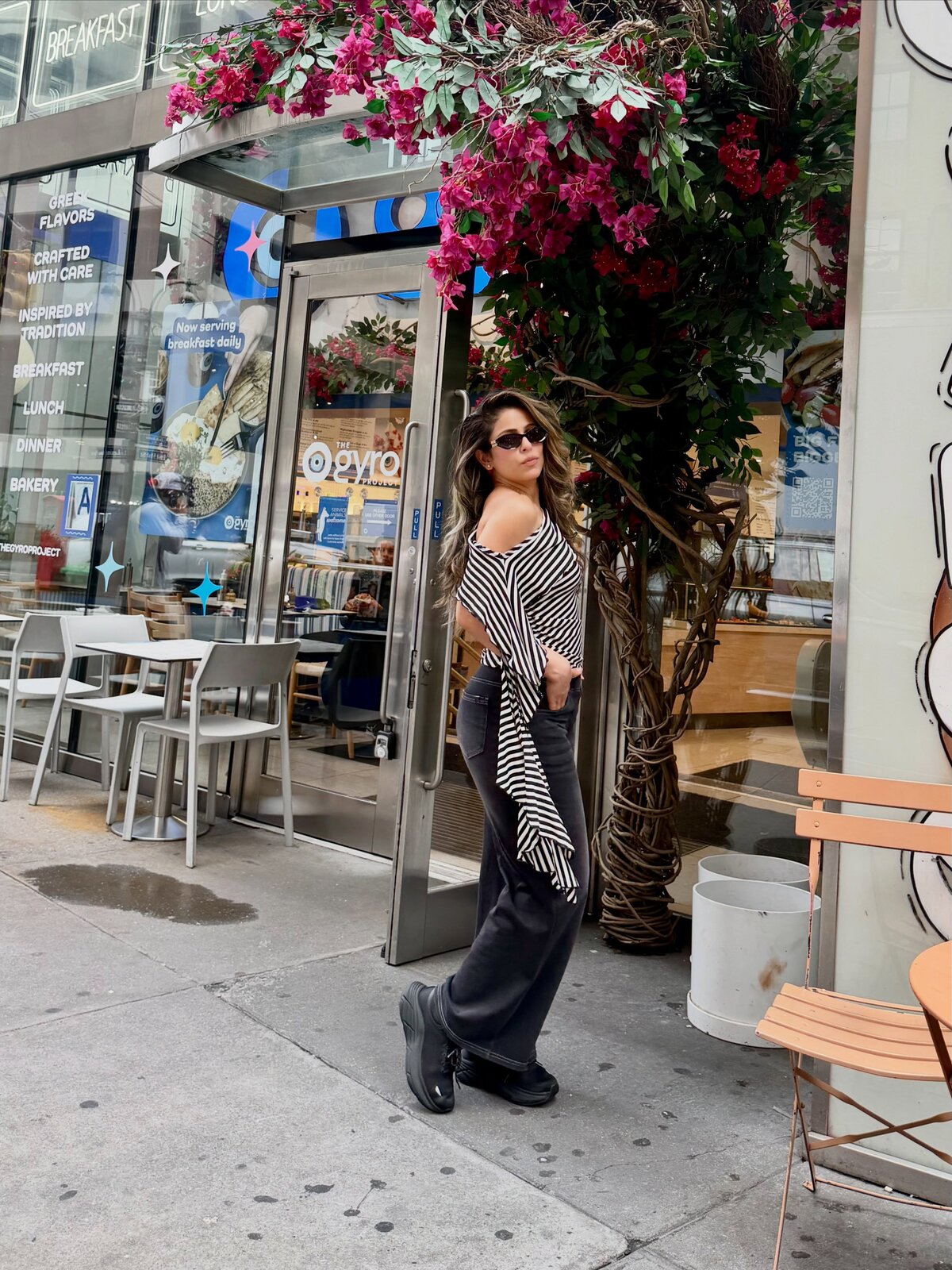 Roopsi Bansal in a black and white striped off-shoulder top and black wide-leg jeans, standing under a bougainvillea-covered awning outside The Gyro Project in New York City