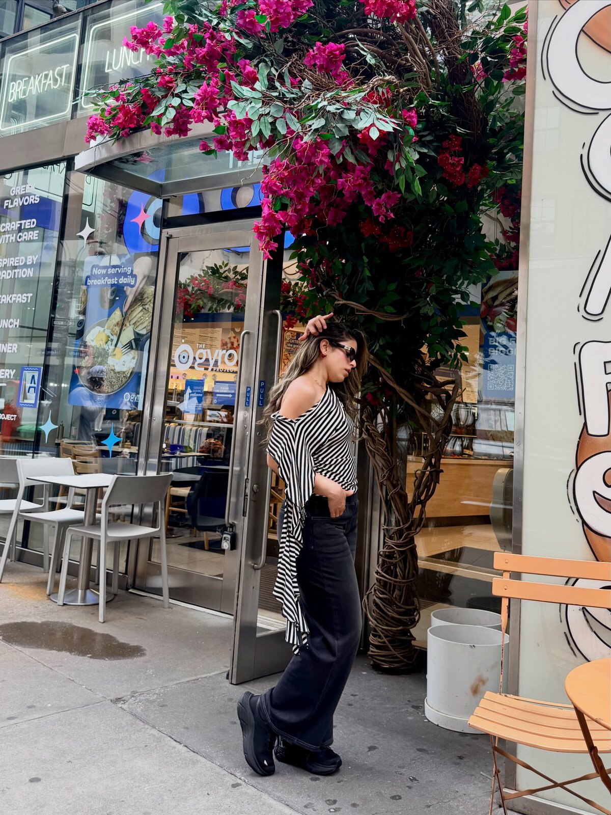 A second angle of the same striped off-shoulder top and black wide-leg jeans outfit, under the same bougainvillea-covered awning