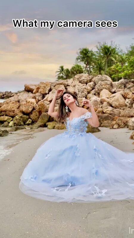 Roopsi Bansal in a sky-blue floral ball gown, posed against a rocky jetty on the beach, the polished camera version of a phone-vs-camera comparison reel